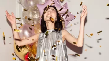 A woman in a shiny dress joyfully throws gold confetti in the air by balloons.