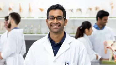 Smiling man in lab coat with glasses in a science classroom.