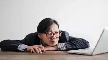 Person with glasses leaning on table, looking at laptop screen thoughtfully