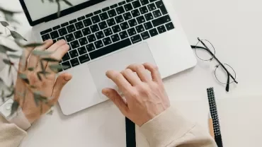 Person working on a laptop at a desk with glasses and a notebook nearby.