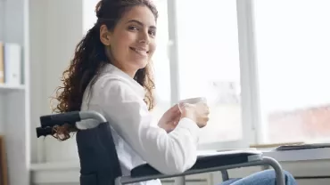 Woman in a wheelchair smiling while holding a cup and sitting by a laptop in a bright room.