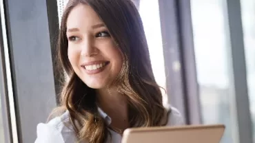 Smiling woman holding a tablet, looking out a window.