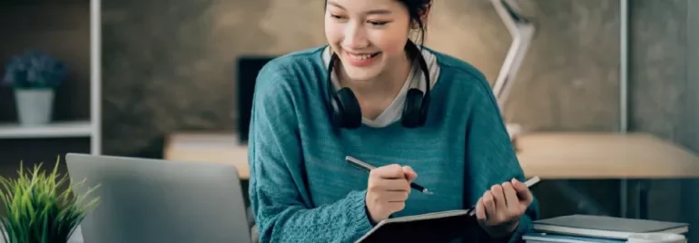 Smiling woman with headphones writing notes at a laptop.
