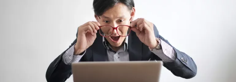 Man looking surprised while holding glasses and staring at a laptop.