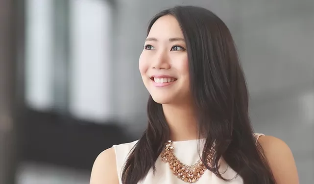 Woman smiling brightly, wearing a white dress with a statement necklace, against a blurred background.